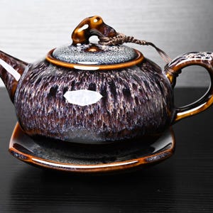 Op de afbeelding: Een keramische theepot met een glanzend, donkerbruin en paars glazuur met een gevlekte, kristallijne textuur. De theepot heeft een gebogen tuit, een handvat en een deksel met een kleine knop die met een bruin koord aan het lichaam is bevestigd.