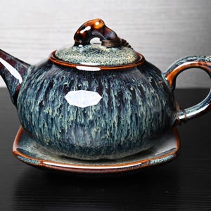 Może przedstawiać: Ceramiczny czajniczek z pasującym kwadratowym spodkiem, pokryty reaktywnym szkliwem w odcieniach głębokiego błękitu, turkusu i brązu. Czajniczek posiada zakrzywioną wylewkę, uchwyt oraz pokrywkę z małym uchwytem, z pionowym wzorem zacieków.