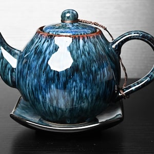 Op de afbeelding: Een keramische theepot met een glanzende, diepblauwe en zwart gestreepte glazuurafwerking. De theepot heeft een gebogen tuit, een bijpassend deksel met een kleine knop en een bruin koord dat het deksel met het handvat verbindt.