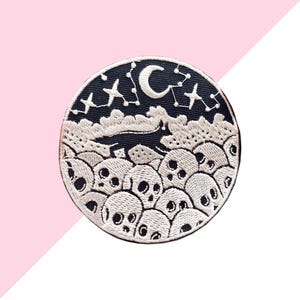 May include: Embroidered patch with a circular design featuring a night sky with a crescent moon and stars. Below the sky are clouds and a collection of skulls. The patch is black, white, and gray.