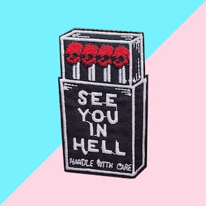 May include: Embroidered patch depicting a black matchbox design with four red skull matches. The text "SEE YOU IN HELL" is in white, with "HANDLE WITH CARE" below. The patch is set against a pink and blue background.