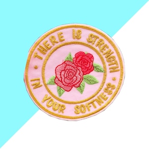 Op de afbeelding: Geborduurde patch met een roze achtergrond, met twee rode rozen met groene bladeren. De patch heeft gouden randen en de tekst "THERE IS STRENGTH IN YOUR SOFTNESS."