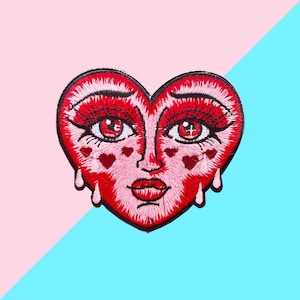 Op de afbeelding: Geborduurde patch in de vorm van een rood hart met een gezicht. Het gezicht heeft rode ogen, lange wimpers en kleine hartvormige accenten. Twee tranen bevinden zich onder de ogen. De patch staat op een roze en blauwe achtergrond.