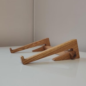 Puede incluir: Un soporte para portátil de madera con un acabado de madera natural. El soporte está hecho de dos piezas de madera que se unen para crear una plataforma elevada para un portátil.