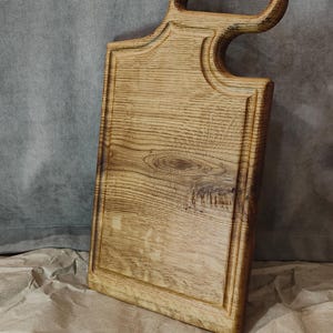 May include: A rectangular wooden cutting board with a handle. The board is made of light-colored wood with visible grain and a dark border. The handle has a unique, curved design. The board is approximately 15 inches long and 8 inches wide.