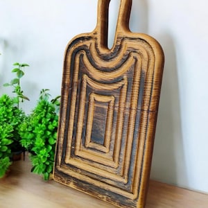 May include: A wooden cutting board with a unique design featuring concentric circles and a handle. The board has a dark brown finish and is made of natural wood.