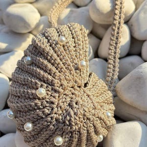May include: A beige, crocheted seashell-shaped bag with a matching strap. The bag is adorned with several faux pearls. The bag is resting on a bed of white stones. The bag's design is reminiscent of nautical themes.