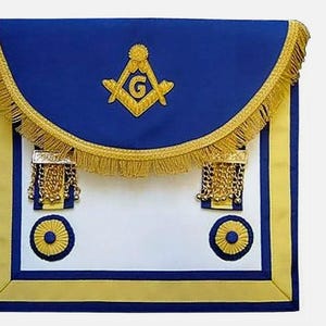Może przedstawiać: Niebieski fartuch masoński z ozdobnymi złotymi lamówkami i haftowanym złotym kompasem i kątownikiem z literą 'G' w środku. Fartuch ma dwa złote frędzle i dwa złote koła z niebieskimi środkami.