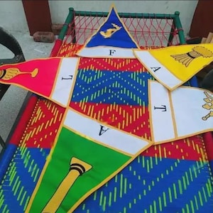 Può includere: Un oggetto decorativo a forma di stella, colorato, con bandierine rosse, gialle, blu e verdi. Ogni bandierina presenta un design unico e le lettere F, L, A, T e V. L'oggetto è posto su un letto a fantasia.