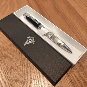 May include: A black and silver pen with intricate designs, presented in a black box with a white interior. The pen features a black top, silver accents, and a detailed engraved pattern on the barrel. The box has a Masonic symbol.