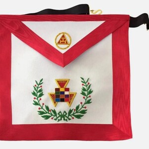 Könnte beinhalten: Eine weiße Freimaurer-Schürze mit rotem Rand und goldener Stickerei. Die Schürze zeigt einen Kranz aus grünen Blättern und ein goldenes und rotes Symbol in der Mitte. Die Schürze hat einen schwarzen Gurt mit einer goldenen Schnalle.
