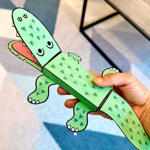 Könnte beinhalten: Ein grünes Papierspielzeug in Form eines Alligators mit einem dreieckigen Muster auf dem Rücken und einem roten Maul. Der Alligator hat schwarze Augen und weiße Zähne. Das Spielzeug wird von einer Hand gehalten.