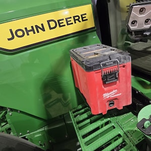 May include: A red Milwaukee Packout tool box sits on a green tractor. The tractor has a yellow and black John Deere logo. The tool box has a black handle and latches. A green circular object with a black handle is also visible.