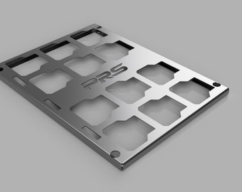 May include: A rectangular, silver-colored metal plate with multiple square cutouts. The letters "PRS" are cut out in the center. The plate has rounded corners and appears to be a component or part.