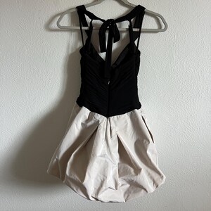 K&ouml;nnte beinhalten: Ein schwarzes und beiges Kleid mit einzigartigem Design. Das Oberteil ist schwarz und gerafft, mit einem V-Ausschnitt und Spaghettitr&auml;gern, die sich im R&uuml;cken zu einer Schleife binden. Der Rock ist beige und hat eine Ballonform.