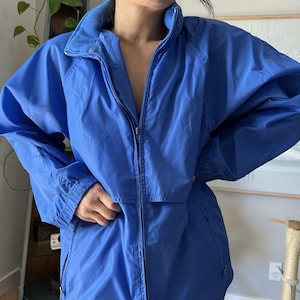 May include: A bright blue windbreaker jacket featuring a high collar, a full-length front zipper, and long sleeves with elasticized cuffs. The jacket has a smooth, lightweight fabric construction with a simple, functional design and side pockets.