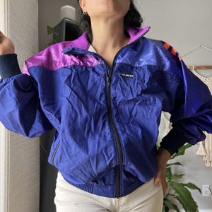 May include: A Louis Garneau windbreaker jacket featuring a color-blocked design in shades of purple, royal blue, and navy. The jacket has a full-length front zipper, long sleeves with dark navy ribbed cuffs, and a high collar. The brand name is printed.