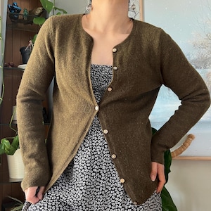 May include: Olive green cardigan with a round neckline and button closure. The long-sleeved sweater is made of a textured material. The cardigan is worn over a black and white floral print dress.