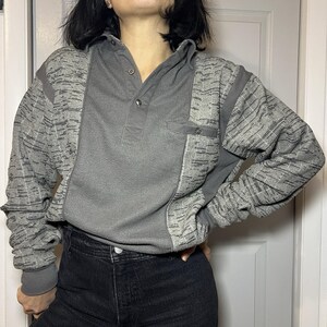 Könnte beinhalten: Ein graues Langarmshirt mit Kragen und einer Knopfleiste mit drei Knöpfen. Die Ärmel und Schulterpartien weisen ein strukturiertes Muster auf, während das Mittelteil eine gerippte Textur hat. Eine kleine Tasche ist auf der Brust sichtbar.