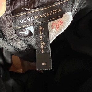 K&ouml;nnte beinhalten: Nahaufnahme eines schwarzen Kleidungsst&uuml;cketiketts mit dem Markennamen BCBGMAXAZRIA in wei&szlig;er Schrift. Ein wei&szlig;es Etikett mit roten Markierungen ist angebracht. Ein kleineres schwarzes Etikett lautet "Made in China" und hat die Nummer "04".