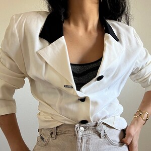 May include: Cream-colored blazer with a black collar and black buttons. The blazer has a double-breasted design and a tailored fit. The sleeves are slightly gathered at the shoulders. The blazer is paired with light-colored denim jeans.