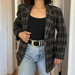 May include: A black and brown plaid blazer with a black tank top and blue jeans. The blazer has a classic plaid pattern in shades of black, brown, and tan. A black belt with a gold buckle cinches the waist.