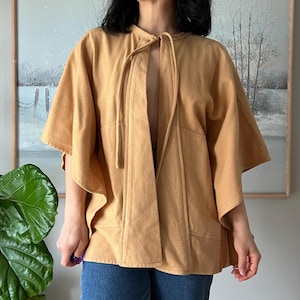 May include: A tan-colored, open-front cape-style jacket with short sleeves. The jacket features a tie closure at the neckline and two front pockets. The garment is made of a soft, textured fabric and is worn over blue jeans.