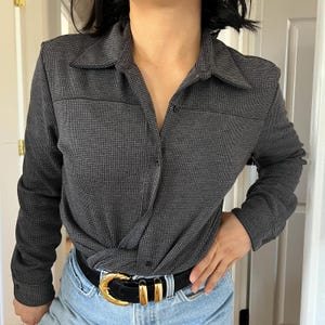 May include: A dark gray button-down shirt with a small houndstooth pattern. The shirt has long sleeves, a collar, and a knot detail at the waist. It is paired with blue jeans and a black belt with a gold buckle.