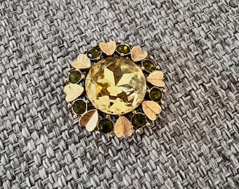 May include: A round, gold-toned brooch with a large, faceted yellow gemstone center. The brooch is encircled by smaller green gemstones, gold-toned leaf shapes, and a gold-toned metal frame. The brooch is set against a gray textured background.