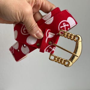 May include: A red fabric belt with white polka dots and anchor designs. The belt has a gold-colored chain buckle. The belt is held in a hand.