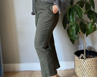 May include: Olive green suede pants with a straight leg cut. The pants have a high waist and pockets. The person is wearing a gray sweater and brown shoes. A potted plant is in the background.