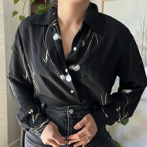 May include: A black long-sleeve button-down shirt featuring a delicate print of thin, tan-colored grass stems and small, fluffy white seed heads scattered across the fabric. The shirt has a classic collar and white buttons down the front placket.