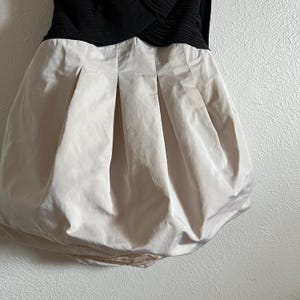K&ouml;nnte beinhalten: Ein schwarzes und beiges Kleid mit einem einzigartigen Design. Der obere Teil ist schwarz mit einem strukturierten, plissierten Aussehen. Der Rock ist beige und hat einen Ballon-Saum und vertikale Falten, die eine volumin&ouml;se Silhouette erzeugen. Das Kleid ist aus gl&auml;nzendem Stoff.