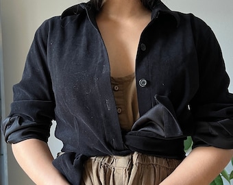 May include: Black button-down shirt with satin cuffs and a collared neckline. The shirt is worn open over a tan top and paired with tan pants. The outfit is a casual, yet stylish ensemble.