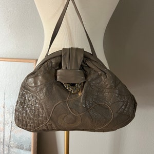 May include: A taupe leather handbag with a top handle. The bag features a woven pattern on the lower left, a crocodile-like texture, and decorative stitching. It has a clasp with a gold-colored metal accent.