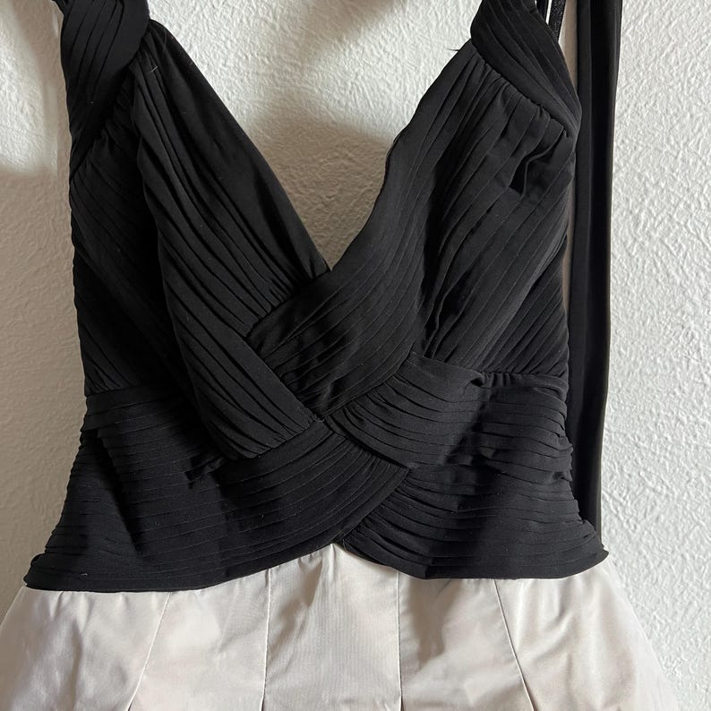 K&ouml;nnte beinhalten: Ein formelles Kleid mit einem schwarzen Oberteil und einem cremefarbenen Rock. Das Oberteil hat ein plissiertes Design mit einem Kreuzdetail an der Brust. Das Kleid hat einen Neckholder-Ausschnitt und einen flie&szlig;enden Rock.