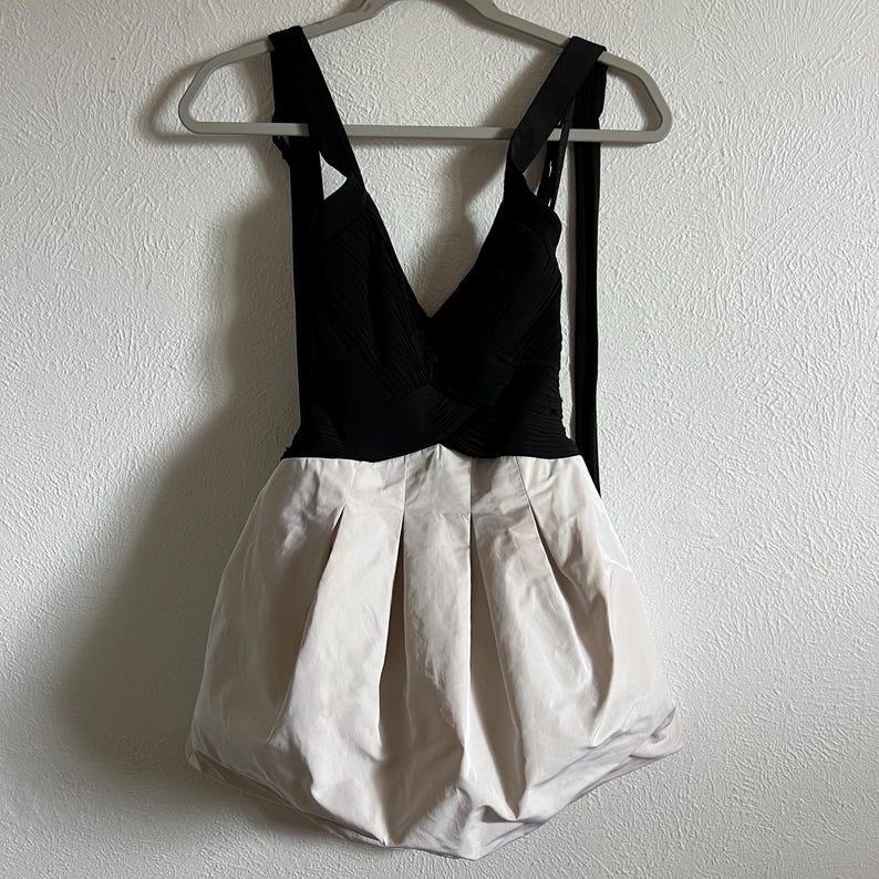 K&ouml;nnte beinhalten: Ein schwarzes und beiges Kleid mit V-Ausschnitt und plissiertem Rock. Das Kleid hat ein eng anliegendes Oberteil und einen ausgestellten Rock, der an einem grauen Kleiderb&uuml;gel h&auml;ngt.