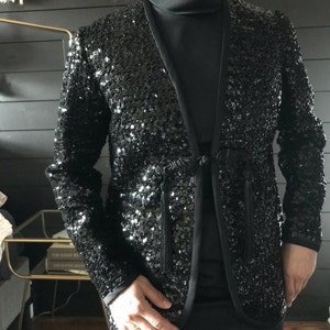 Könnte beinhalten: Eine schwarze Paillettenjacke mit einem langen Quasten-Bindeverschluss. Die Jacke hat einen Stehkragen und lange Ärmel.