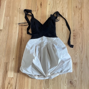 K&ouml;nnte beinhalten: Ein schwarzes und cremefarbenes Kleid mit V-Ausschnitt und gerafftem Rock. Das Oberteil ist schwarz mit einem Kreuzmuster, und der Rock ist cremefarben mit Falten. Das Kleid hat schwarze Tr&auml;ger zum Binden.