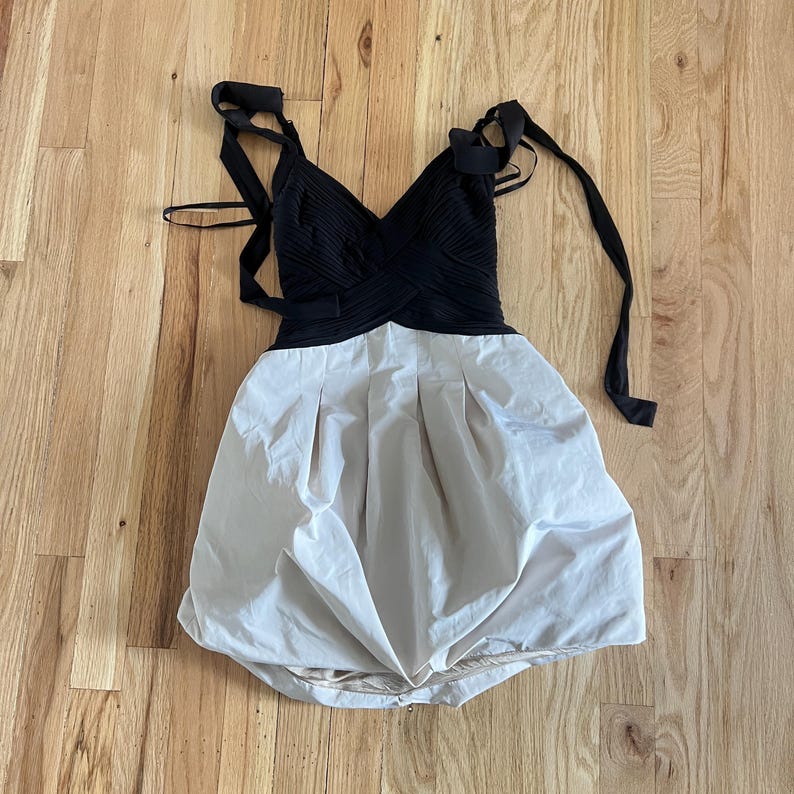 K&ouml;nnte beinhalten: Ein schwarzes und cremefarbenes Kleid mit V-Ausschnitt und gerafftem Rock. Das Oberteil ist schwarz mit einem Kreuzmuster, und der Rock ist cremefarben mit Falten. Das Kleid hat schwarze Tr&auml;ger zum Binden.