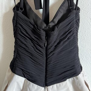 K&ouml;nnte beinhalten: Ein formelles Kleid mit einem schwarzen, gerafften Oberteil und einem hellbeigen Rock. Das Kleid hat einen Neckholder-Ausschnitt und einen Rei&szlig;verschluss hinten. Der Stoff hat ein strukturiertes Aussehen.