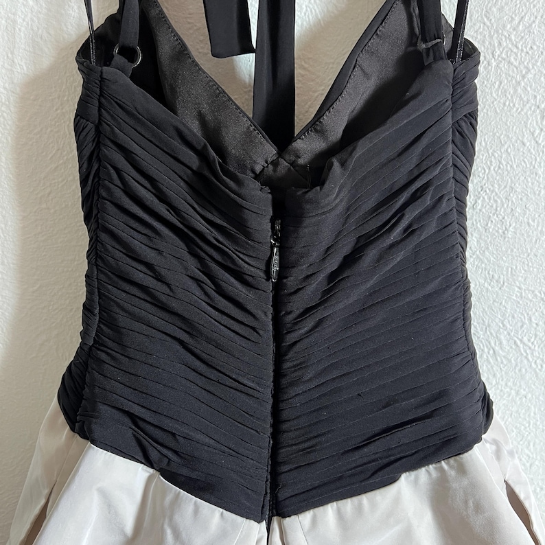 K&ouml;nnte beinhalten: Ein formelles Kleid mit einem schwarzen, gerafften Oberteil und einem hellbeigen Rock. Das Kleid hat einen Neckholder-Ausschnitt und einen Rei&szlig;verschluss hinten. Der Stoff hat ein strukturiertes Aussehen.