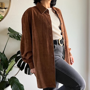 May include: A long, button-down brown suede shirt with a collared neckline. The shirt is worn over a cream-colored ribbed sweater and paired with gray jeans and a black belt with a gold buckle. The sleeves are long and loose.