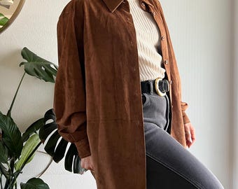 May include: A long, button-down brown suede shirt with a collared neckline. The shirt is worn over a cream-colored ribbed sweater and paired with gray jeans and a black belt with a gold buckle. The sleeves are long and loose.