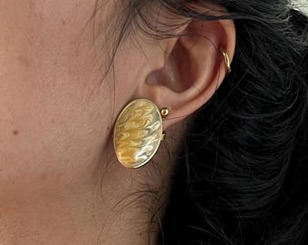 May include: Gold-toned oval clip-on earring with a cream and yellow swirled enamel design. The earring is attached to the earlobe. The earring has a gold-colored frame.