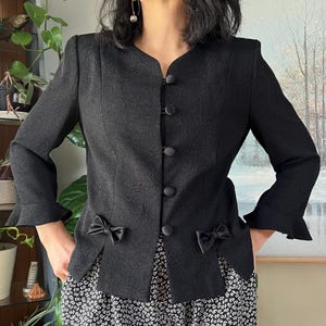 May include: Black blazer with a textured floral pattern, featuring a V-neckline and button closure. The blazer has small bows at the waist and flared sleeves. The garment is paired with a floral print skirt.