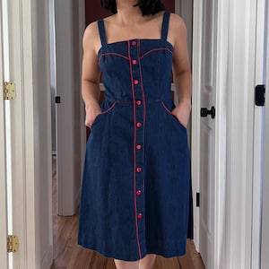 Könnte beinhalten: Dunkelblaues Jeanskleid mit roter Paspelierung und Knöpfen. Das Kleid hat einen quadratischen Ausschnitt, eine Knopfleiste vorne und Taschen. Das Kleid hat eine Midi-Länge und ein figurbetontes Oberteil.