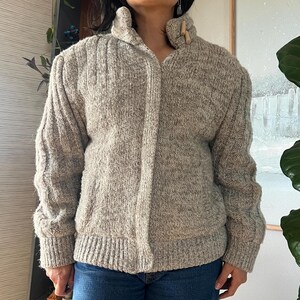 May include: A light brown, textured knit cardigan with a high collar and wooden toggle closure. The sweater has a ribbed design on the sleeves and a fitted waist. The overall style is a cozy, vintage look.