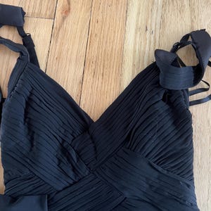 K&ouml;nnte beinhalten: Ein schwarzes Abendkleid mit V-Ausschnitt und plissiertem Oberteil. Das Kleid hat Spaghettitr&auml;ger und eine Schleife. Der Stoff scheint ein flie&szlig;endes Material zu sein, das sich f&uuml;r besondere Anl&auml;sse eignet.