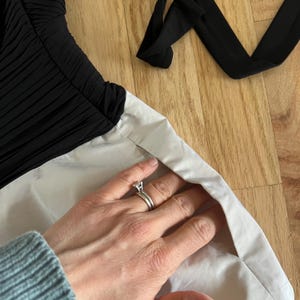 K&ouml;nnte beinhalten: Ein schwarz-wei&szlig;es Kleid mit einem plissierten Oberteil und einem hellfarbigen Rock. Ein schwarzer Riemen ist im Hintergrund zu sehen. Das Kleid liegt auf einer Holzoberfl&auml;che.